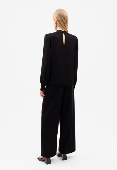 Schwarze Bluse mit langen Ärmeln und Schlüssel-Ausschnitt auf der Rückseite, kombiniert mit weiten schwarzen Hosen. Glattes, leichtes Material.