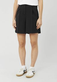 Shorts plissés noirs avec une coupe ajust�ée et une ceinture, fabriqués en tissu lisse. Assortis à des baskets blanches et des chaussettes de cheville.