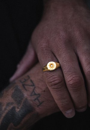 Tume nahaga käsi kannab kuldsõrmust, millel on päikesekiirte kujundus ja väike keskmine kalliskivi, toetudes tätoveeritud käsivarrel.