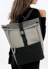 Grauer und schwarzer Rucksack mit Roll-Top-Design, aus wasserabweisendem Material. Verfügt über einen Schnallenverschluss und eine mit Reißverschluss versehene Tasche.