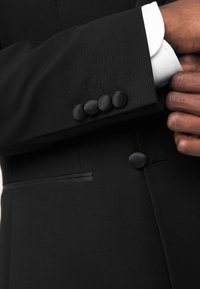 Black blazer sleeve featuring five textured buttons, a slim fit, and contrasting white cuff. Smooth fabric with clean lines and subtle stitching.