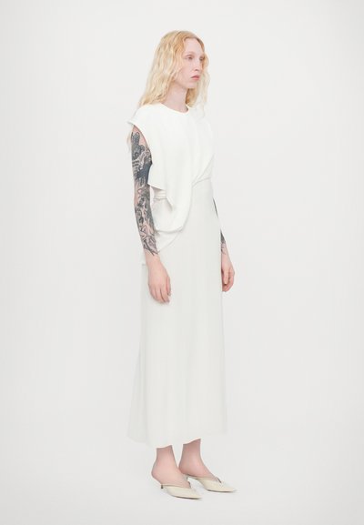 Weißes ärmelloses Kleid mit drapiertem Frontdetail, bodenlang, aus glattem Stoff. Komplementiert durch spitze weiße Absätze.