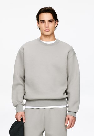 Młody mężczyzna w jasnoszarej bluzie i pasujących spodniach, trzyma czarną czapkę w lewej ręce, neutralne tło.