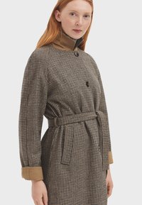 Abrigo de espiga marrón con cuello beige, cierre de botones, cintura ceñida con cinturón y mangas largas con acentos en los puños. Tela texturizada.