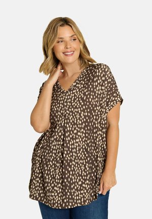 Mujer sonriente con cabello rubio hasta los hombros que lleva una blusa marrón suelta con escote en V y manchas beige, y vaqueros azules sobre un fondo liso.