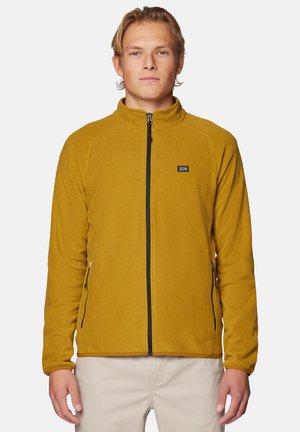 Junger Mann trägt eine senfgelbe Fleecejacke mit schwarzem Reißverschluss und beigen Hosen, steht vor einem schlichten hellen Hintergrund.