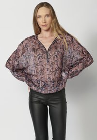 Durchsichtige Bluse mit langen Ärmeln im Paisleymuster in Mauve, Blau und Weiß. V-Ausschnitt mit Reißverschlussdetails. Kombiniert mit schwarzen, figurbetonten Hosen.