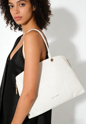 Femme aux cheveux bouclés portant un haut noir sans manches et un grand sac cabas blanc Pierre Cardin sur l'épaule.