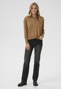 Brun stickad tröja med krage och fyra knappar, ihopparad med mörka jeans och svarta skor. Tyget ser mjukt och texturerat ut.