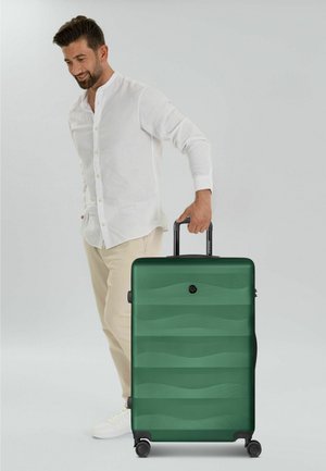 Hombre con camisa blanca y pantalones beige de pie, sosteniendo el asa de una gran maleta rígida verde con cuatro ruedas.