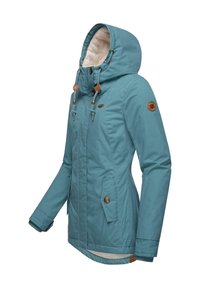 Veste d'hiver turquoise avec une capuche bordée de fourrure, une fermeture éclair frontale, des poches avec boutons-pression et des accessoires décoratifs en métal. Présente un ourlet arrière allongé.