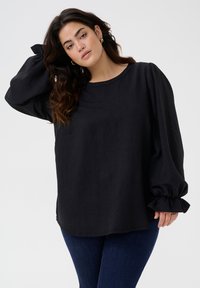 Black, long-sleeve blouse with a round neckline and textured fabric. Features gathered sleeves with elastic cuffs. Paired with blue denim.