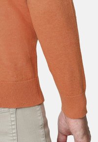 Suéter de punto naranja con dobladillo y puños de canalé. Textura ligera, diseño ajustado, exhibido en una persona con pantalones color crema.
