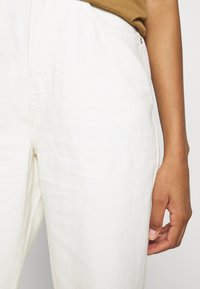 Calças de linho brancas com uma textura subtil, apresentando uma cintura de altura média e bolsos laterais. Uma blusa na cor oliva parcialmente visível acima.