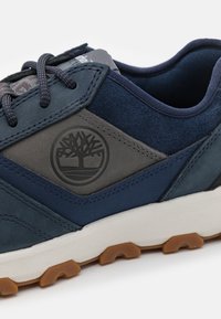 Sötétkék és szürke sneaker, texturált velúrbőr felsőrésszel, gumitalppal és domború logóval. Tűzött díszítések és kerek fűzők jellemzik.