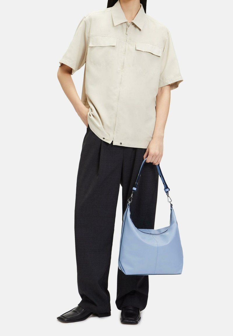 Personne portant une chemise beige à manches courtes et un pantalon noir à jambes larges tenant un sac à main bleu clair avec une bandoulière.