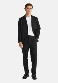 Melnā uzvalkā no gludas auduma ar vienrindu žaketi un taisnajām biksēm, kombinēts ar baltu T-kreklu un melnām kurpēm.