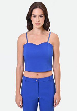 Femme aux longs cheveux foncés porte un top camisole court bleu vif et un pantalon taille haute assorti avec fermeture à boutons, debout sur un fond blanc.