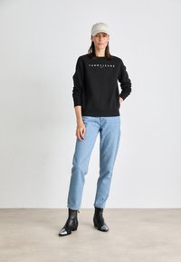 Felpa nera con logo "TOMMY JEANS", abbinata a jeans azzurri chiari e stivaletti neri con punta in argento. Un cappellino neutro completa il look.