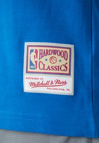 Niebieska bawełniana koszulka z białą prostokątną metką z kolorowym logo "Hardwood Classics", emblemą NBA oraz napisem "Mitchell & Ness".