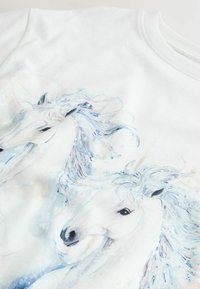 Weißes Stoff-T-Shirt mit einem Aquarelldruck von zwei weißen Einhörnern mit blauen Akzenten, die fließende Mähnen und ausdrucksstarke Merkmale betonen.