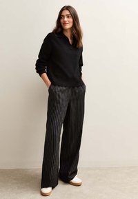 Czarny sweter z kołnierzykiem, w parze z szerokimi spodniami w czarno-białe paski. Modelka nosi jasne buty, stoi z dłońmi w kieszeniach.