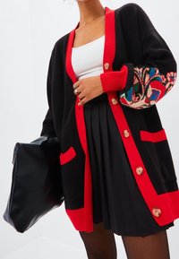 Cardigan nero con dettagli rossi, caratterizzato da un ricamo floreale colorato sulle maniche, chiusura con bottoni e due tasche frontali.