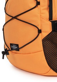 Pomarańczowy plecak z teksturowanego materiału, czarną siateczką i elastycznym designem sznurka. Zawiera logo Eastpak na materiałowej naszywce.