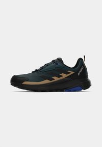 Trail running-skor med en robust design i svart och mörkgrön, texturerad ovandel och bruna detaljer samt en blå yttersula för bra grepp.