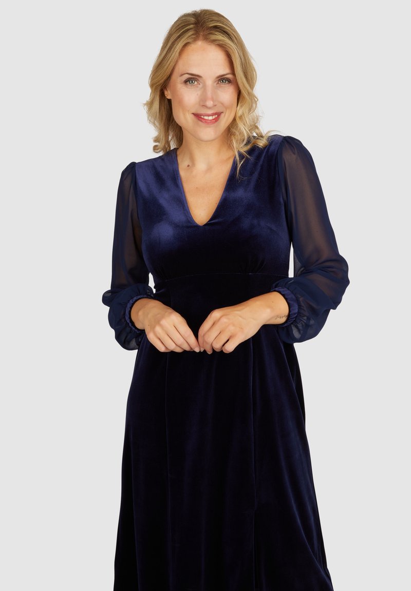 Navy Samt Kleid mit langen durchsichtigen Ärmeln, einem V-Ausschnitt und gerafften Manschetten. Der Stoff hat eine glatte Textur und einen leichten Glanz.
