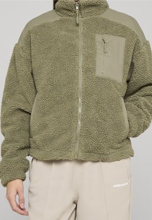 Persoon draagt een olijfgroene fleecejas met rits en borstzak en beige broek met het Pegador-logo op de dij.