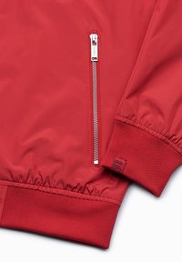 Manche de veste rouge avec poignet côtelé et détail de poche zippée en métal sur tissu lisse.