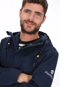 Marineblauw waterdicht jack met een ritssluiting aan de voorkant, trekkoorden voor een verstelbare capuchon en een logo op de linker mouw. Getextureerd materiaal en een gladde afwerking.