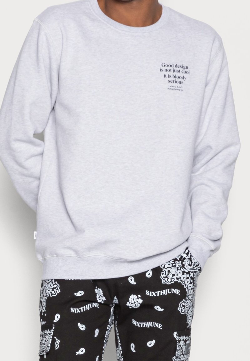 Sudadera gris de cuello redondo con texto en el lado izquierdo del pecho. Pantalones negros con un patrón de paisley blanco y el nombre de la marca repetido a lo largo.