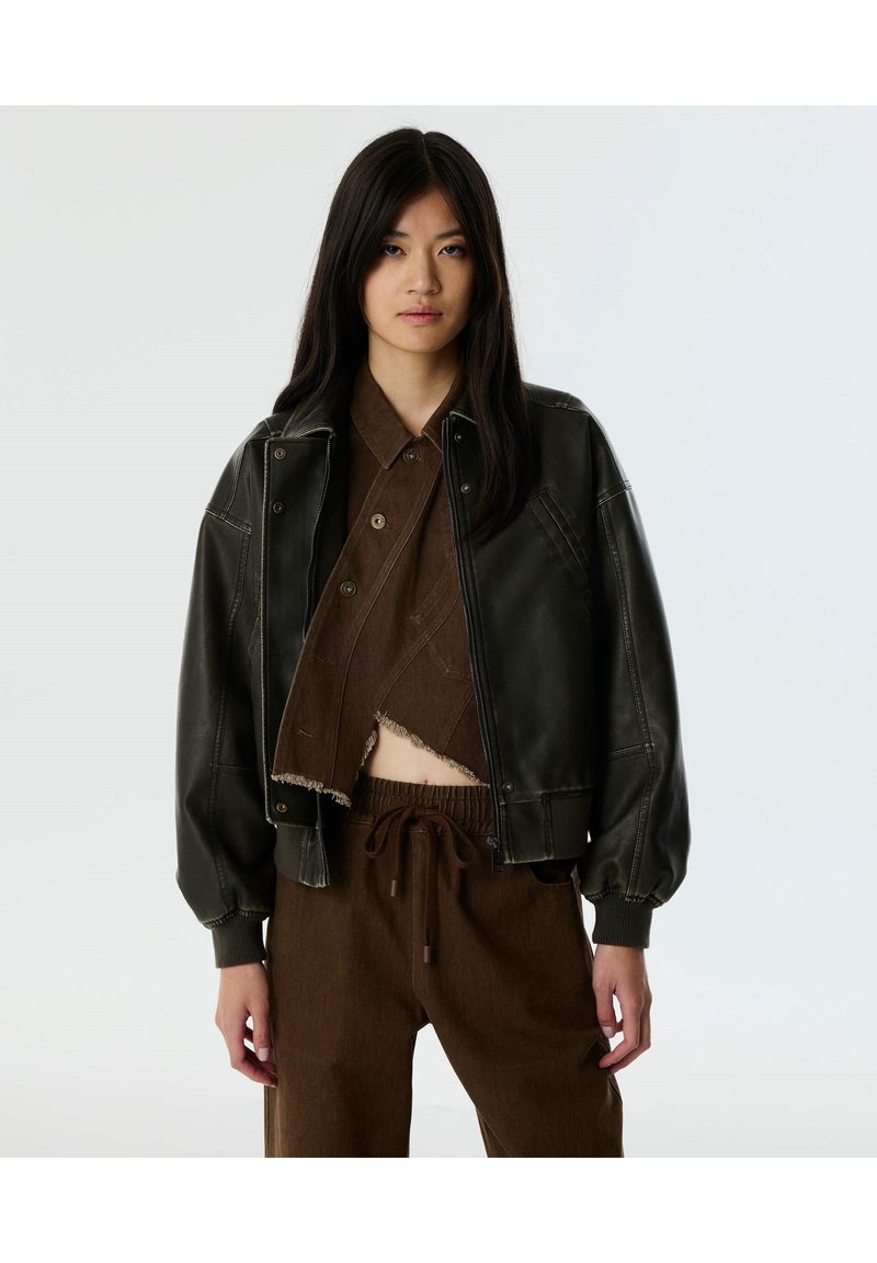 Chaqueta de cuero negra recortada con puños acanalados, ajuste holgado, costuras en contraste, llevada sobre una camisa recortada marrón con botones y pantalones a juego.