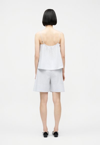 Γυναίκα με κοντά μαύρα μαλλιά, που φοράει αμάνικο τοπ με ανοιχτό μπλε και λευκές ρίγες και αντίστοιχα σορτς μέχρι το γόνατο, στέκεται με την πλάτη προς τα εμπρός.