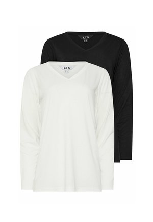 Blondynka stoi oparta o ścianę, ubrana w białą bluzkę z długim rękawem i dekoltem w serek, czarne spodnie oraz białe trampki z gumowymi podeszwami.