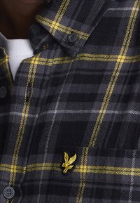 Camisa de cuadros en negro y gris con acentos amarillos, cuello abotonado y logotipo bordado en el bolsillo del pecho. Tela texturizada con un patrón a cuadros.
