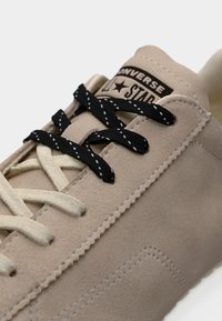 Μπεζ αθλητικό παπούτσι Converse με κορδόνια σε μπεζ και μαύρο μοτίβο, ορατή ραφή και ετικέτα "Converse All Star" στη γλώσσα.