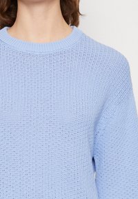 Detailní záběr osoby v světle modrém pleteném svetru s vroubkovaným kulatým výstřihem a detailním texturovaným vzorem.