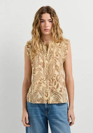 Jonge vrouw met golvend haar, die een beige mouwloze blouse met knopen en een bruine bladprint draagt, en een blauwe spijkerbroek, staand tegen een effen achtergrond.