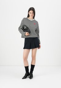 Maglione a righe bianco e nero, maniche lunghe, vestibilità ampia, logo a forma di cuore rosso. Gonna mini plissettata nera, calze alla caviglia nere, scarpe a punta.