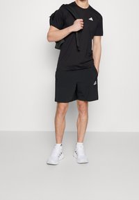 Svart atletisk t-shirt och shorts med vitt Adidas-logga; ihop med vita sportskor. Texturen verkar jämn och åtsittande.