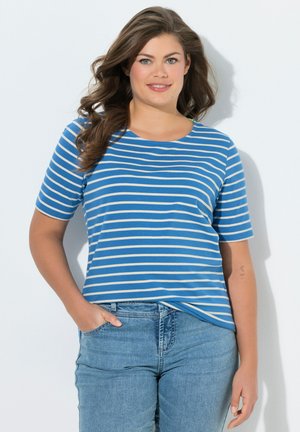 Vrouw met lang bruin haar, een blauw-wit gestreept shirt met korte mouwen en lichtblauwe spijkerbroek, staand met één hand in de zak.