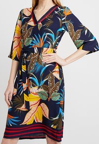 Blommig klänning med marinblå bakgrund, V-ringning, trekvarts ärmar med klockform och ett midjebälte med randig nederdel.