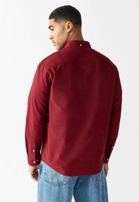 Chemise bordeaux à manches longues avec col à boutons, tissu texturé et poignets à un seul bouton, associée à un jean en denim bleu clair.