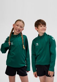 Grønne sportsjakker med halv lynlås lavet af glat stof, med logo og kontrasterende hvide detaljer på skuldrene. Båret med sorte shorts.