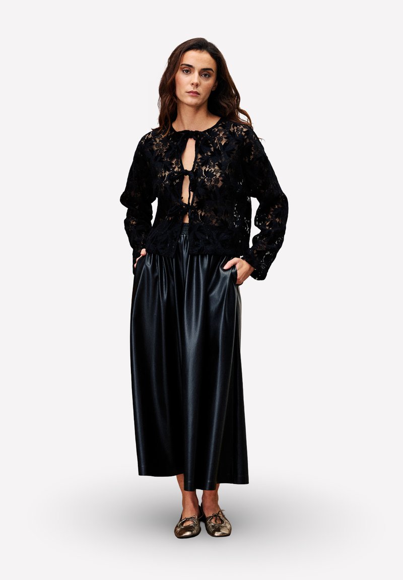 Frau in einem langen schwarzen Lederrock und einem durchsichtigen schwarzen Spitzentop mit Bändern, steht mit den Händen in den Taschen und trägt goldene Ballettschuhe.