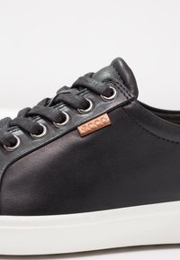 Schwarze Ledersneaker mit strukturiertem Obermaterial, flachen grauen Schnürsenkeln, silbernen Ösen und einer hellgrauen Gummisohle; mit einem kleinen braunen Logostreifen.