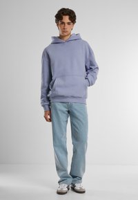 Lys lilla hettegenser laget av mykt stoff, med en frontlomme og justerbar hette, kombinert med lyseblå jeans og hvite joggesko.